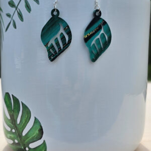 Marbled green acrylic leaf earrings hanging on a white ceramic planter with painted tropical leaves.