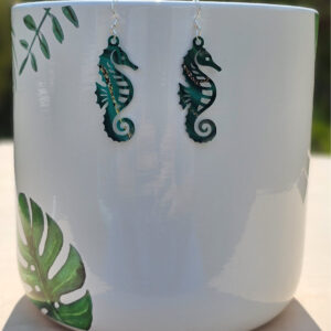 Seahorse-shaped acrylic earrings in deep green and gold marbled pattern hanging on a white planter with tropical leaf design.