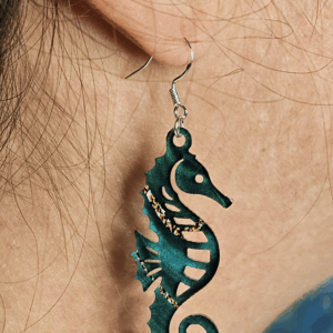 Laser-cut acrylic seahorse earring in deep teal with gold accents worn on ear.