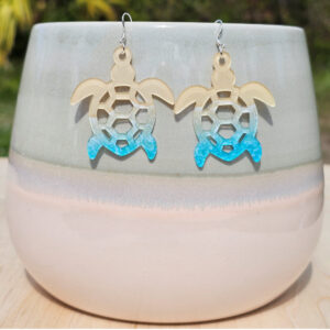 Sea turtle earrings in tan and blue gradient acrylic hanging on a pastel ceramic planter outdoors.